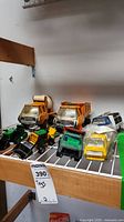 Seven vintage toy vehicles and parts displayed on a white wire rack, including a green loader, orange cement mixer truck, yellow dump truck, and white van with Pepsi decal. Toys show use wear and scratches.