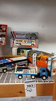 Photo of several toy vehicles including Greyhound buses in and out of packaging, a Pepsi truck with bottles, and a Kenner truck in box placed on a wooden surface with a label.