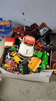 Box filled with assorted vintage toy vehicles including metal trucks, cars, and a Hot Wheels car in box