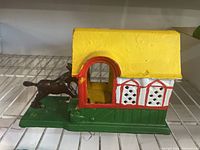 Side view of cast iron kicking mule bank showing the mule on the left side and a small house with a yellow roof, red and white walls on the right.