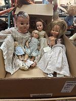 Six vintage dolls arranged sitting and lying in cardboard box, showcasing size variety and clothing.