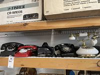 Four rotary dial phones on a wooden shelf including a small red toy phone, two black rotary phones, and a white and brass decorative phone.