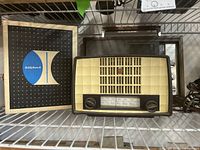 Vintage radio with cream front panel, two black control knobs, tuning dial with numerical markers and red emblem, dark brown casing placed on metal shelving with Bell & Howell box nearby.
