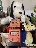 Photo showing a collection of Snoopy-themed toys including a plush, a radio toy, and a music box stand around various other toys.