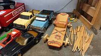 Wide view showing black plastic Chevrolet Corvette toy car, blue metal dump truck, yellow metal Tonka truck, painted wooden toy car, and wooden paddles.