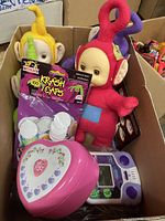 Box containing three Teletubbies plush dolls, Krash Caps game, pink heart-shaped case, and Goosebumps handheld game