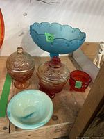 Photo of blue compote dish, two covered candy dishes (amber and reddish-orange), small reddish-orange candy dish, and pale blue Avon saucer dish on wooden surface