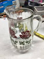 Clear ribbed glass pitcher decorated with red flowers, greenery, and white picket fence motif. Glass lid with spout placed on top of pitcher.