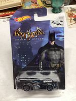 Photo of unopened Hot Wheels 2014 Batman Arkham Asylum Batmobile die-cast car in packaging showing front side with Batman character artwork and logos.