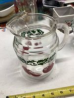 Front angled view of clear glass pitcher with red fruit and green leaf design, showing handle, body decoration and spout.