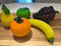 Five art glass fruits arranged on a wooden surface: pear, green apple, orange, banana, grapes clustered.