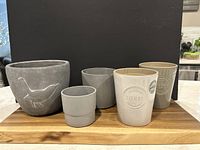 Set of five outdoor plant pots on a wooden board against black background: one large cement pot with bird motif, two gray IKEA pots, two ceramic 'Herbs' planters