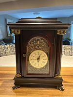Front view of the dark wood mantle clock showing the clock face with Roman numerals and decorative gold accents.