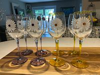 Eight crystal wine glasses displayed on wood surface, four with yellow stems and four with purple stems, clear bowls with daisy designs.