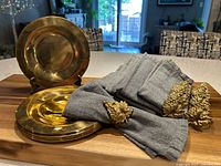 Full lot overview showing brass plates stacked and displayed vertically, gray napkins, and brass napkin holders arranged on a wooden surface.