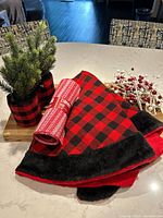 Red and black plaid Christmas tree skirt with faux fur trim, cloth ribbon roll with red and white pattern, and decorative berry strand on a kitchen counter.