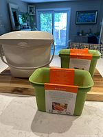 Photo showing one white City of Ottawa compost bin with built-in handle and vented lid, alongside two green freezer compost bins with informational labels.