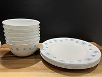 Stack of six white Corelle bowls with blue snowflake pattern beside six matching 10.5" dinner plates on wood surface with black background.