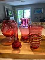 Four cranberry glass vases displayed together on a wooden surface showing different shapes and designs in varying translucent ruby red color.
