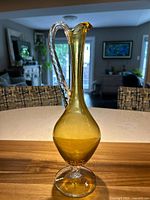 Full view of yellow Empoli glass decanter with elongated neck and clear twisted handle on round base, set on table with background of room.