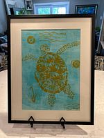 Full view of framed fabric art depicting a gold sea turtle on turquoise background with black frame and beige mat.