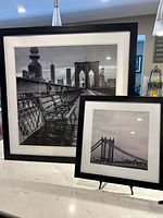 Full view of both framed black and white photographs showing Brooklyn Bridge bench scene and Manhattan Bridge with Empire State Building, on a kitchen counter.