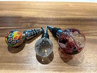 Set of three glass art wine stoppers displayed on wood surface, showing diverse colors and designs.