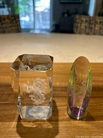 Front view of two glass dragon figurines on table, one rectangular clear glass paperweight with internal 3D laser etched twisting dragon, the other a clear glass prism with rainbow effects and etched dragon and symbol.