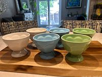 Front angled view of all six stoneware ice cream cups arranged on a wooden tray, showing two white, two light blue, and two green cups with pedestal bases and scalloped bowl shape.
