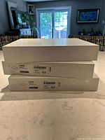 Three white IKEA LACK shelves stacked, two still factory wrapped, one unwrapped, on a white marble countertop with a living room in background.