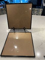 Two black-edged LP record display frames with clear fronts and brown backing, placed on a white countertop. One frame standing on display holders, the other laying flat.