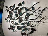 Full view of metal wall art showing the branch and leaf design with mixed brown and turquoise patina finish on a light surface.