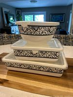 Stacked view of the 3 porcelain containers with decorative black scroll pattern and plastic lids on top, including round bowl and two rectangular containers underneath.