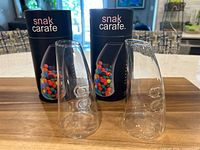Two Snak Carafe glass snack dispensers displayed upright with original boxes behind them on a wood kitchen counter.