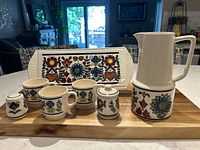 Full view of the complete 7-piece ceramic kitchen set featuring a rectangular cake plate, pitcher, three cups, sugar bowl with lid, and shaker, all with matching colorful floral and heart patterns on white ceramic.