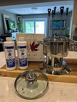 Photo showing the fondue pot with lid, two bottles of fondue fuel, and packaging box in the background on a wooden surface.