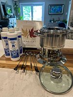 Complete stainless steel fondue set with pot, base, skewers, fuel bottles, and box, shown on wood table in kitchen.