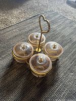Four-piece condiment server set with clear ribbed glass bowls with lids and gold metal holders, arranged on a gold metal tray with heart-shaped handle, placed on carpet.
