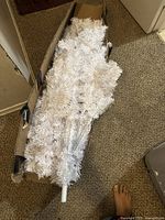 Photo showing the white synthetic Christmas tree disassembled into sections inside a cardboard box, showing the tree's dense white branches and the central white metal pole.