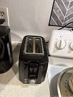 Photo showing front and top of the black Cuisinart 2-slot toaster on a kitchen counter next to an outlet, displaying wear and stains
