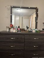 Front view of dark wood six-drawer dresser with metal handles and large framed mirror above it with light bulbs on frame. Personal care items on dresser top.