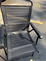Front view showing black folding chair with mesh seat and backrest and black plastic armrests on a paved surface with yellow painted lines