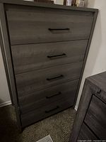 Gray wooden chest of drawers with five drawers and black metal handles shown from front angle against a white wall and carpeted floor.