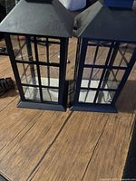 Front view of two black decorative lanterns side by side on a wooden surface showing overall shape and glass panels.