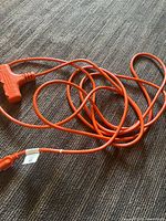 Full view of the bright orange extension cord laid out on a floor showing the T-shaped end with three outlets and the cord.