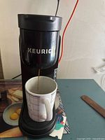 Photo showing Keurig coffee maker brewing coffee into a white mug, black exterior with KEURIG branding.