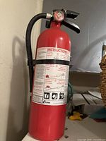 Red Kidde dry chemical fire extinguisher showing front label, attached black hose, pressure gauge, and pin secured with a yellow tie. Photographed in a utility room corner.