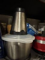 Stacked kitchen appliance components including the motor housing, container bowl with attached blade, and plastic lid stored on shelf.