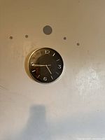Wall clock hanging on a wall. Clock has a black textured face with silver frame and clear Arabic numerals. Some wall marks visible around it.