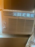 Front view of the white Midea window air conditioner installed in a window, showing two manual control knobs on the left and the Midea logo above the front grille vents.